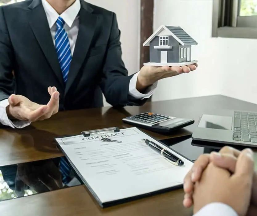 A person in a suit holds a small model house, with a contract, pen, calculator, and laptop on the desk between two people—highlighting key property management steps during important real estate transactions.